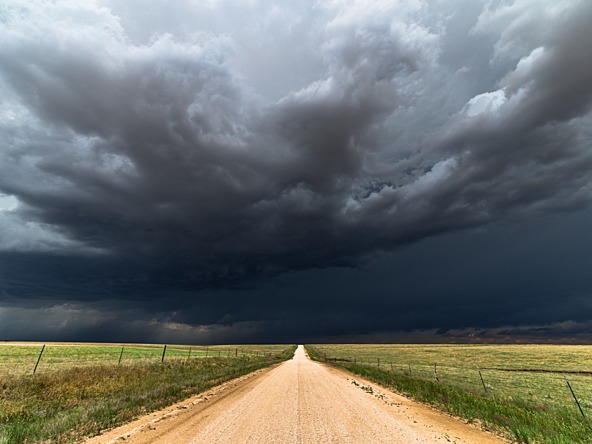 Storm clouds over a long straight road ahead, flanked by fields 